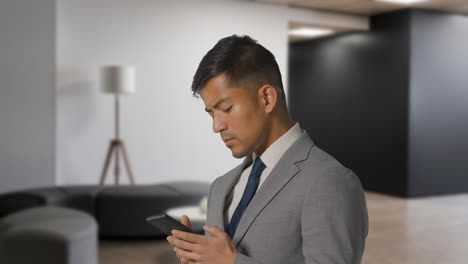 Serious-Businessman-In-Suit-Messaging-On-Mobile-Phone-In-Modern-Open-Plan-Office